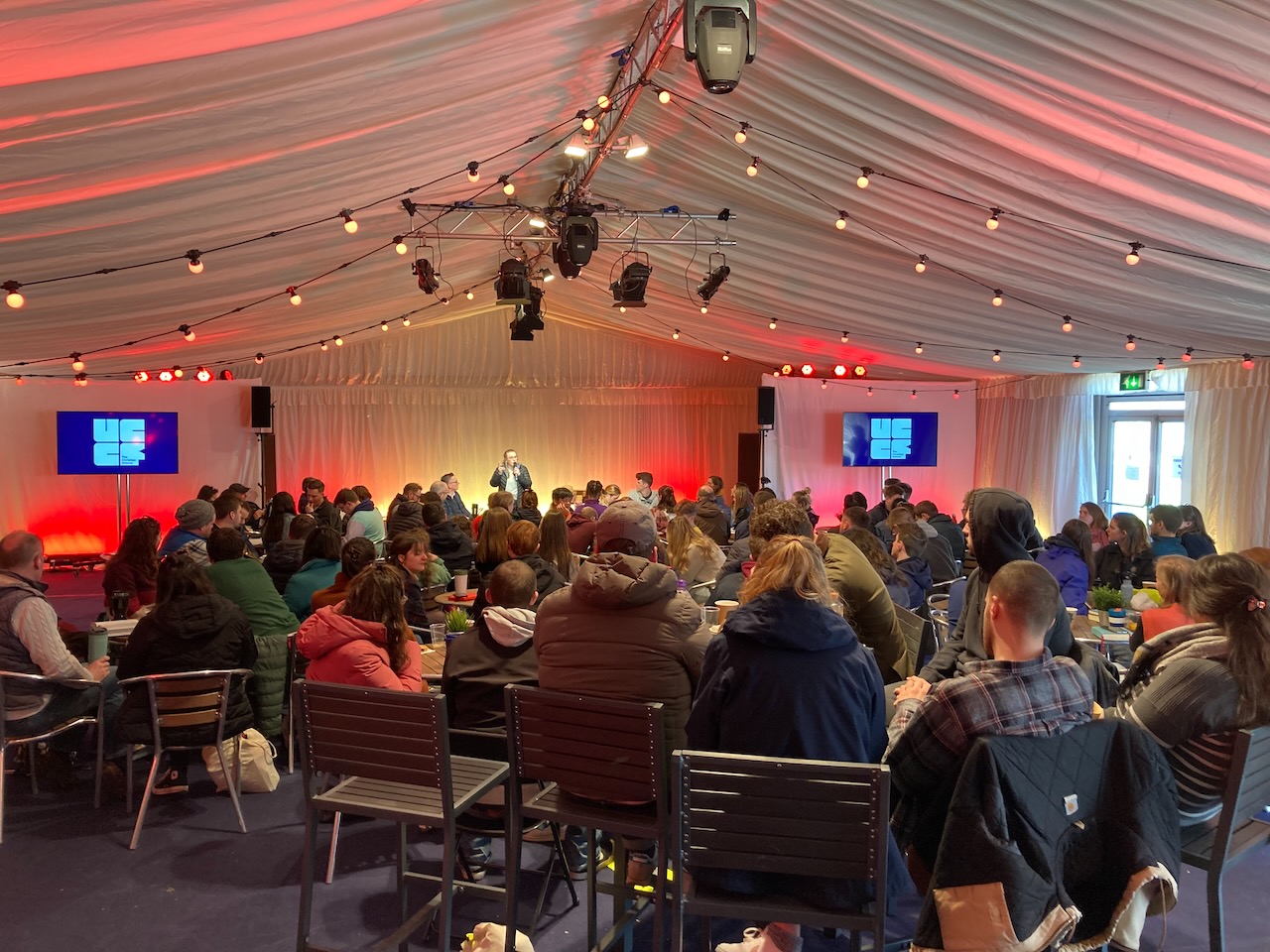 A seminar in the daytime, a speaker is on stage and an audience is listening. The room is lit in red and orange.