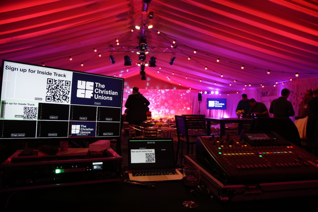 A video switcher, laptop and sound mixer. Red and purple lighting with white star patterns on the stage.
