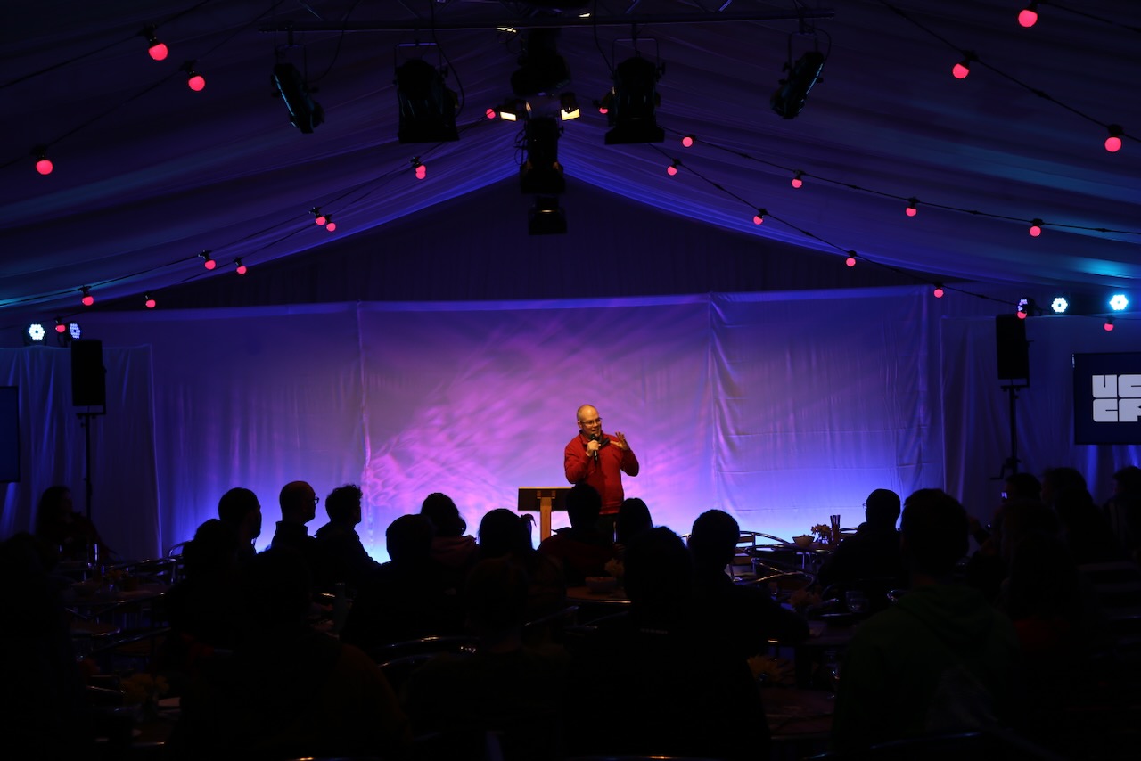 A presenter gives a talk in a darkly lit room with stage lighting
